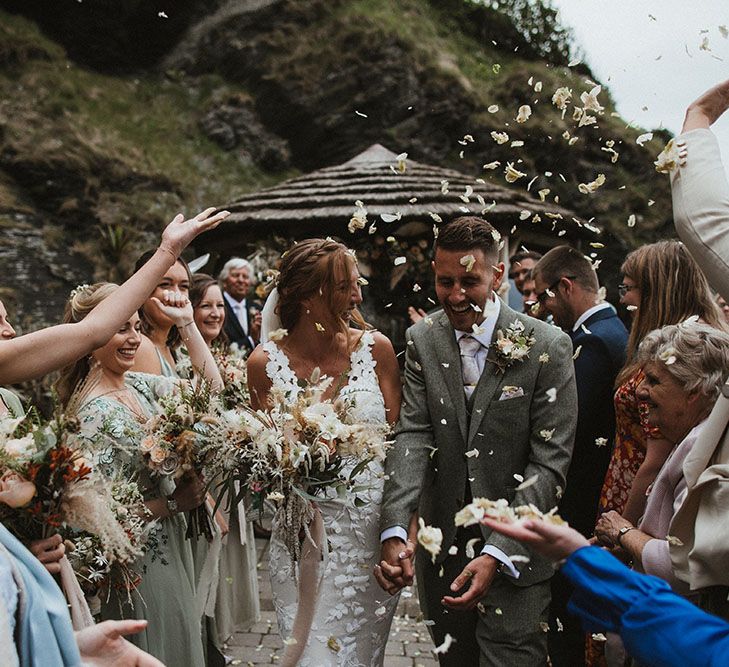 Bride wears lace embellished wedding dress whilst walk through confetti petals with her groom after outdoor wedding ceremony