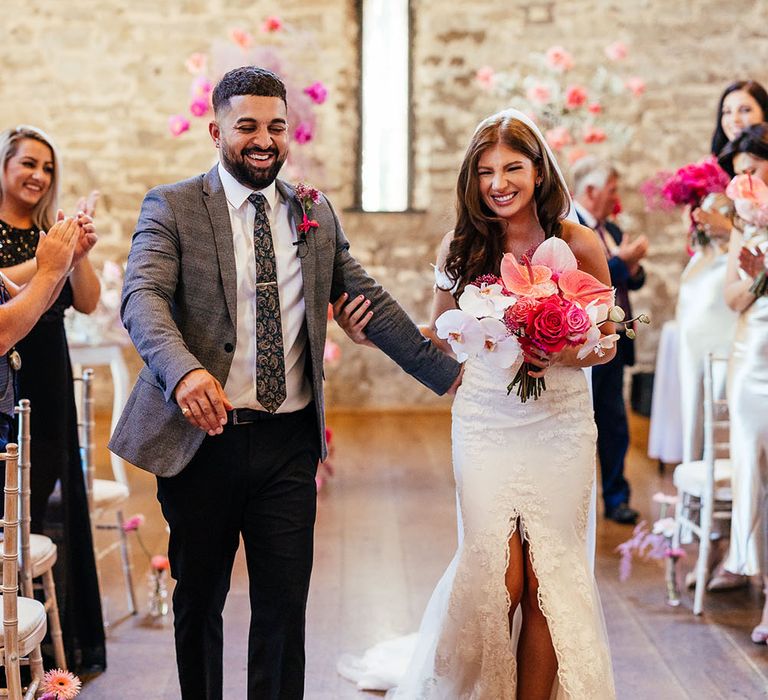 Bride carrying bright pink floral bouquet walks down the aisle with her groom after wedding ceremony at the Priston Mill