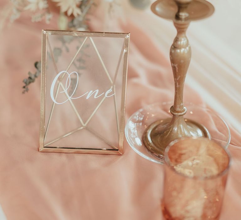 Pink fabric table runner complete with gold and glass frame table numbers next to white candles in gold holders 