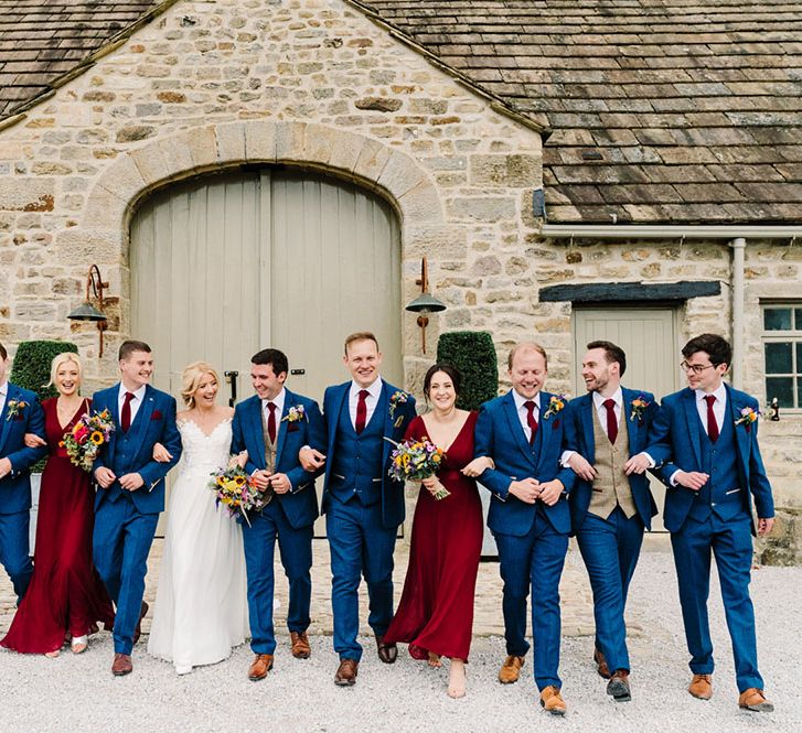 Bride & groom stand with their bridesmaids who wear red bridesmaid dresses with short sleeves and groomsmen in three piece suits 