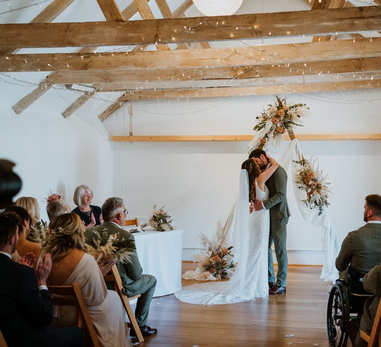 Bride in sleeveless Made With Love Bridal dress with white embellishments and deep plunge back with matching hair ribbon kissing groom in grey suit at The Green Cornwall with triangular wooden arch decoration