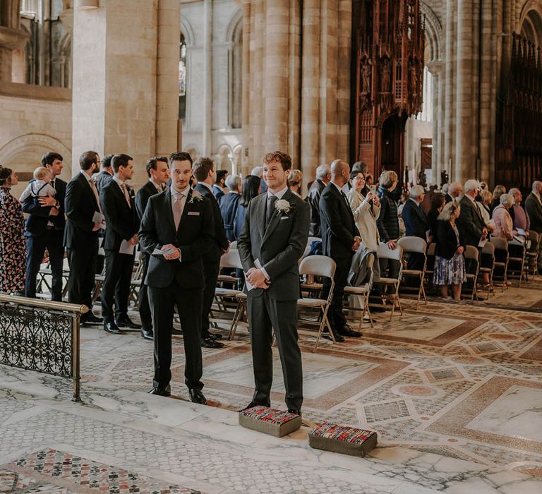 Groom in a dark grey suit with a white rose buttonhole stands with the best man at the altar waiting for the bride 