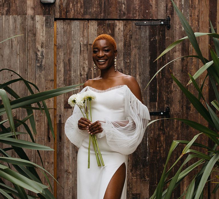 Bride wearing chic off shoulder wedding dress with dramatic puff lace sleeves and nude open-toe heels holding a bunch of white dahlias at 100 Barrington