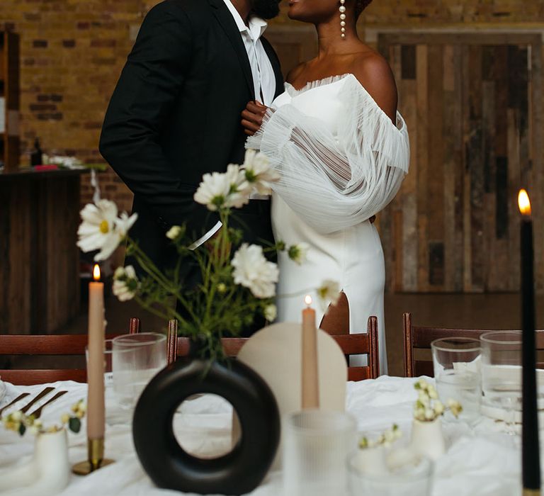 Bride and groom embracing at 100 Barrington standing in front of minimalistic wedding tablescape with black and white circular vases