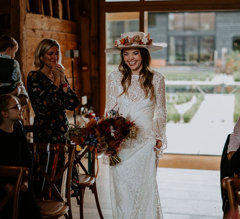 Bride in a laser cut lace wedding dress with a colourful bouquet and white hat with flowers walking down the aisle 