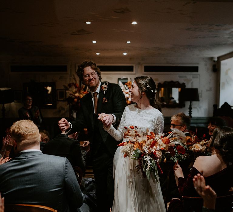 Bride in long sleeve lace vintage wedding dress and closed toe satin heels and groom in dark suit with orange tie dancing down the aisle at The Zetter Townhouse