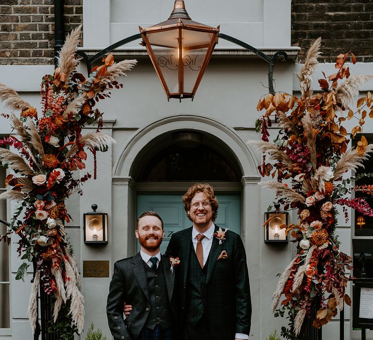 Groom in dark suit with orange tie and autumnal boutonniere standing with best man wearing dark suit, waistcoat, autumnal boutonniere and dark blue tartan kilt underneath large floral arch with roses, peonies, foliage and pampas grass