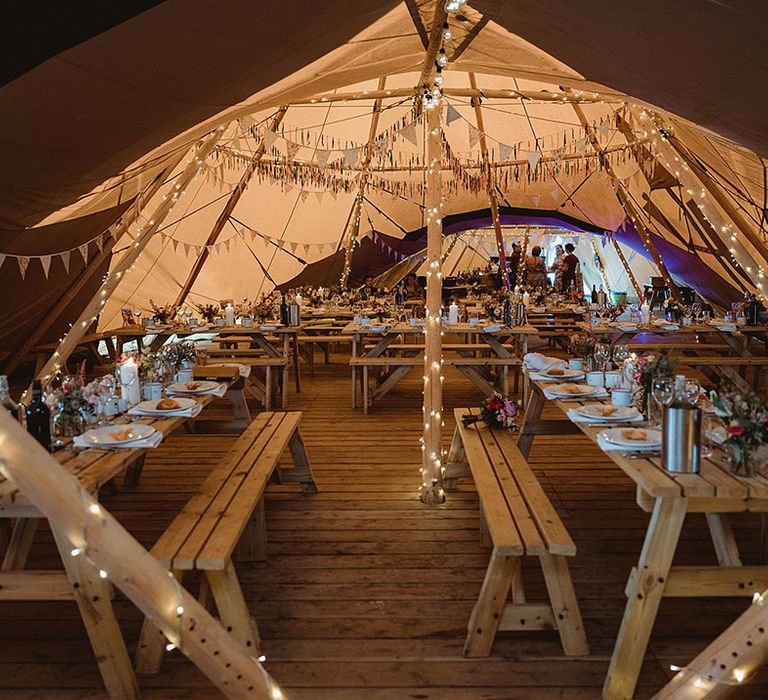 Tipi wedding reception with fairy lights, DIY streamer decor and wooden benches for festival style lesbian wedding 