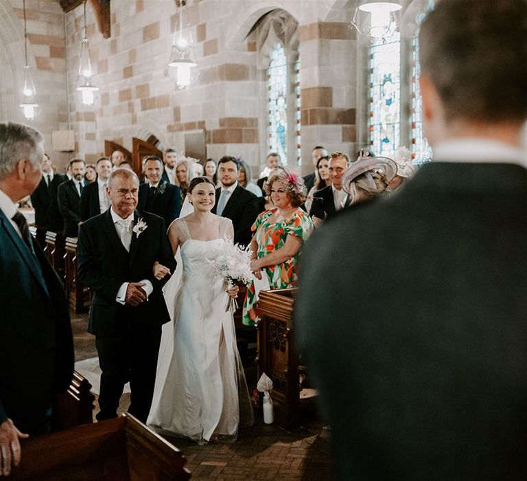 Father of the bride in dark suit with paisley silver tie walks the bride down the aisle for church ceremony 