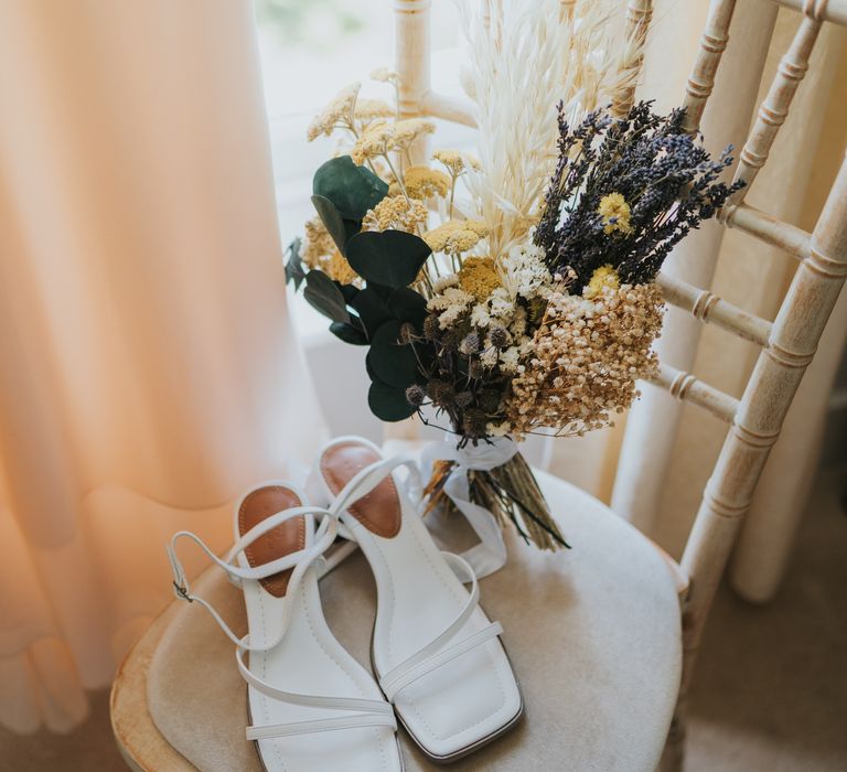 Strappy open-toe heels on cream coloured chair next to dried flower bouquet with white bunny tails, green foliage, yellow dried gypsophila, lavender and baby’s-breath 