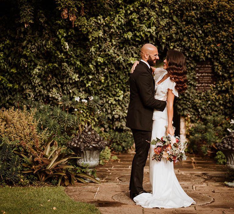 Groom in classic black tuxedo with black bowtie embracing bride in v-neck satin wedding dress with sheer butterfly sleeves and ruffled layer on top holding wedding bouquet with white and pink carnations, garden roses, red strawflower, eucalyptus, baby’s-breath and various dried flowers