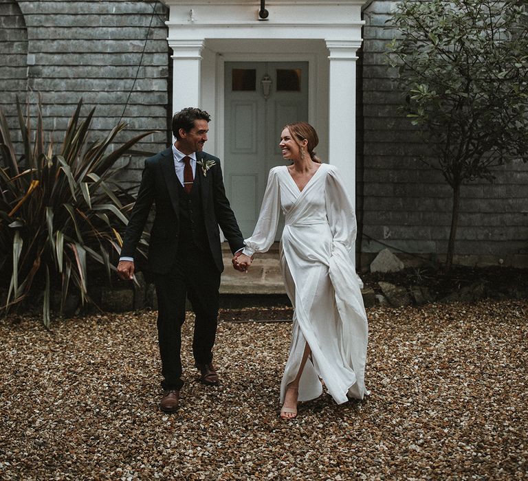 Bride in floaty wedding dress with wrap styling walks alongside her groom in three piece suit and maroon tie 