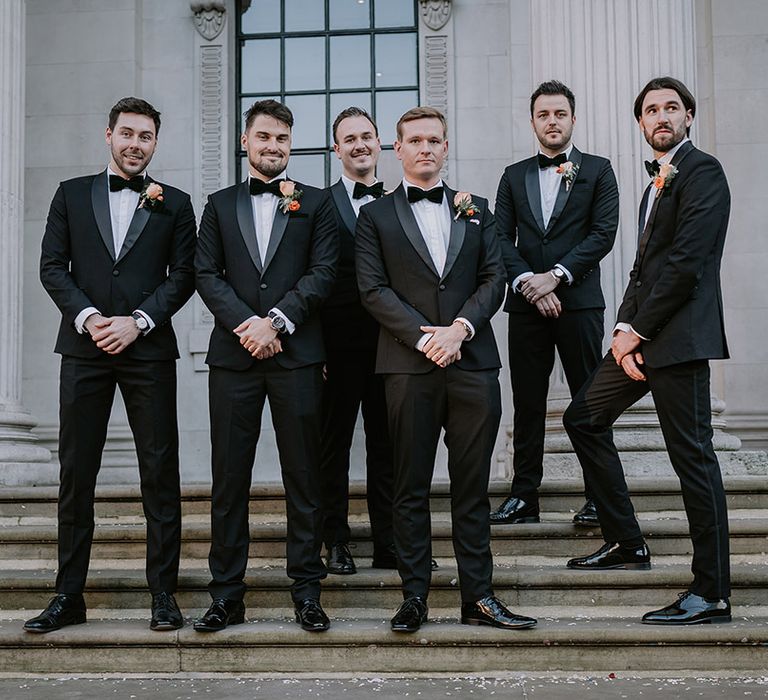 Groom in stylish black tuxedo with black bowtie and light orange rose boutonniere standing with groomsmen in matching black tuxedos 