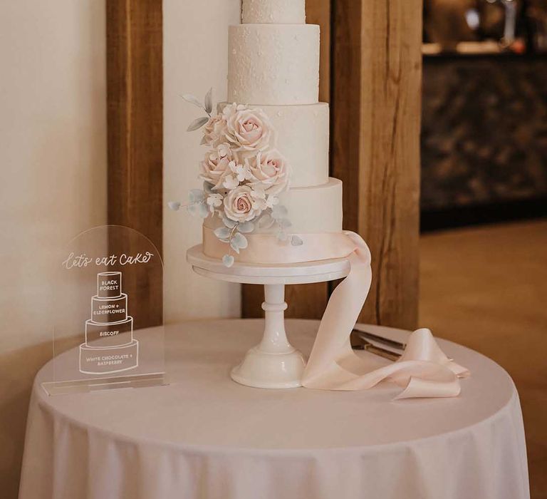 Four tiered buttercream wedding cake with light pink rose and foliage decorations and blush pink ribbon with "let's eat cake" opaque sign