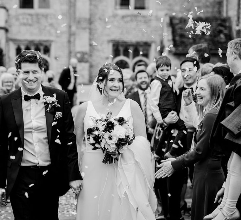 Groom in traditional black tie walking with the bride in a classic Maggie Sottero wedding dress as they have confetti thrown over her 