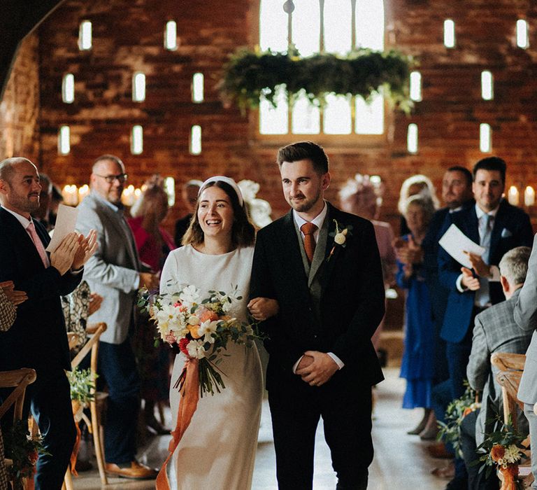 Bride in high neck wedding dress and headbands with wildflower bouquet tied with orange ribbon and groom in three piece suit and grey waistcoat with orange tie for September wedding 