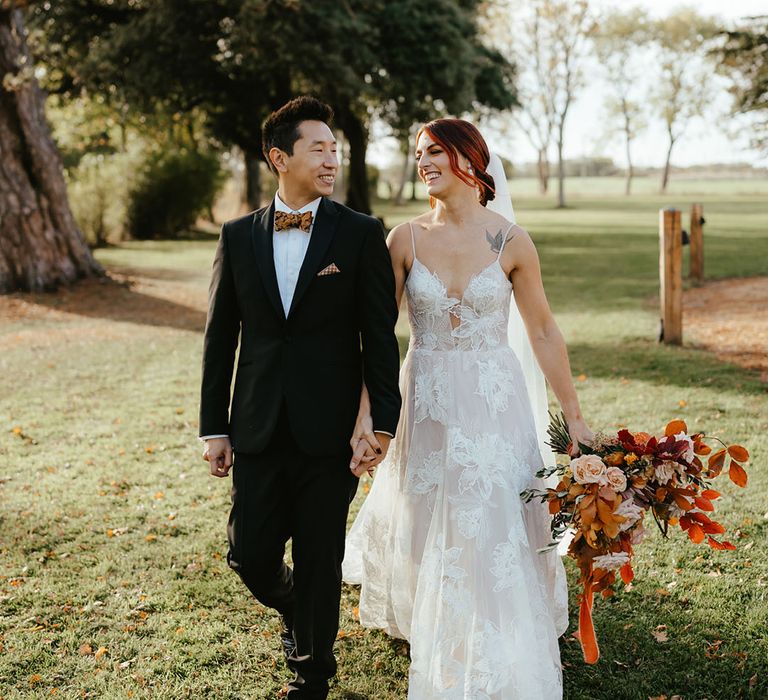 Bride in Madi Lane lace wedding dress holds burnt orange bridal bouquet and walks alongside her groom in black tie 