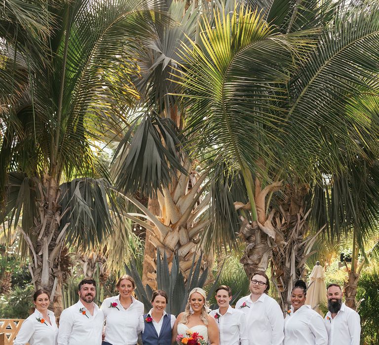 Brides stand with their wedding party who wear blue trousers and white shirts 