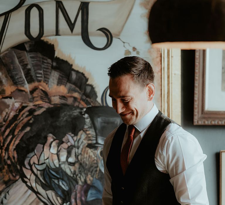 The groom smiles in a black trouser and waistcoat with a white shirt and an orange tie 