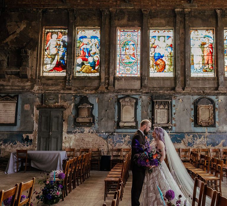 Bride in leaf appliqué sheer Evie Young bridal gown and cathedral length veil holding purple peacock coloured bouquet standing with groom in textured mocha brown three piece grooms suit at London Asylum Chapel 