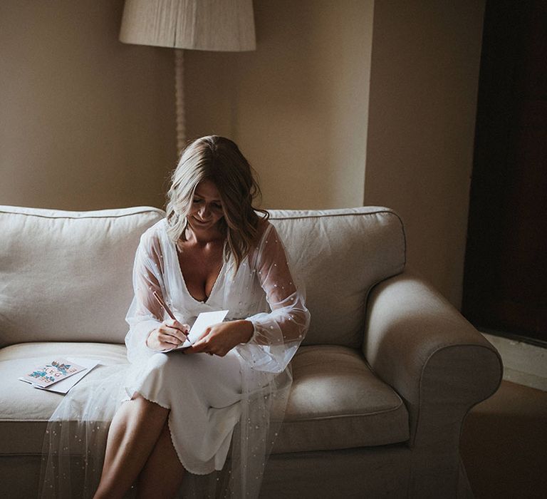 Bride wears pearl embellished sheer robe whilst reading card on the morning of her wedding day