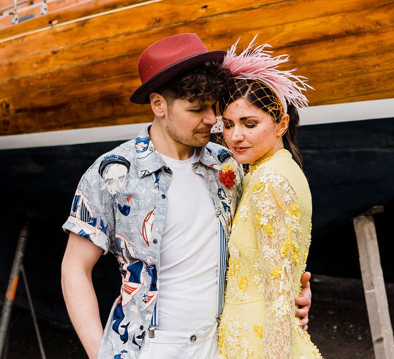 Groom in blue patterned shirt wearing a red hat with the bride in a yellow dress and pink and orange birdcage veil 