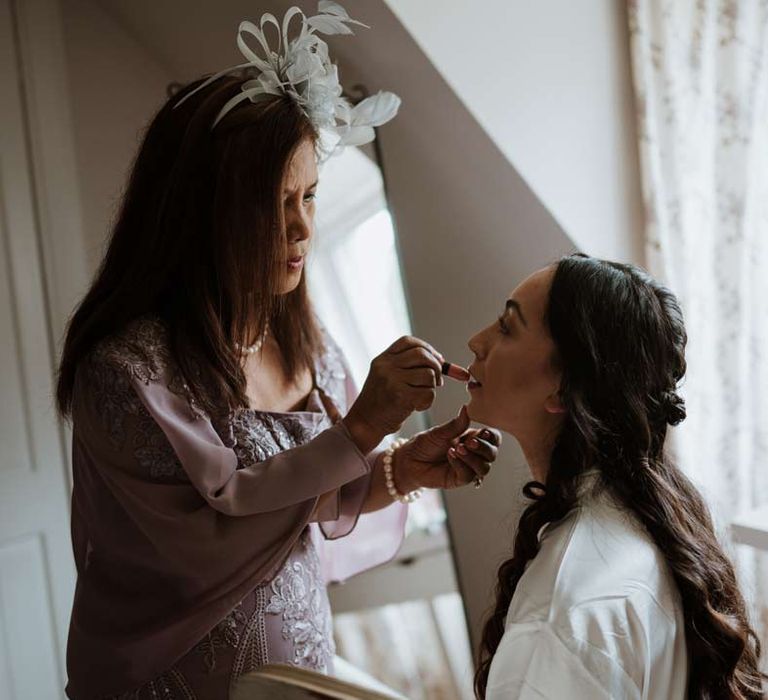 Bride in white satin getting ready robe having her wedding makeup done by wedding guest wearing lilac dress, lilac coverup and white wedding fascinator 