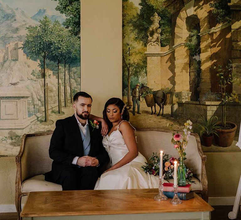 Bride in bow front sleeveless satin wedding dress with large gold bridal hair clip sitting with groom in black blazer with pink garden rose and foliage boutonniere, cornflower blue waistcoat on chaise lounge at Braxted Park wedding venue 