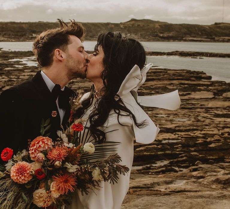 Bride wearing large white hair bow accessory kissing the groom at coastal lighthouse wedding in the North East 