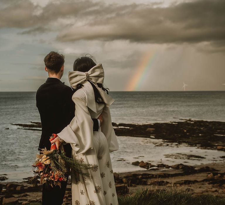 Bride in puff sleeve Bowen Dryden jumpsuit with large white hair bow with groom looking out at the view 