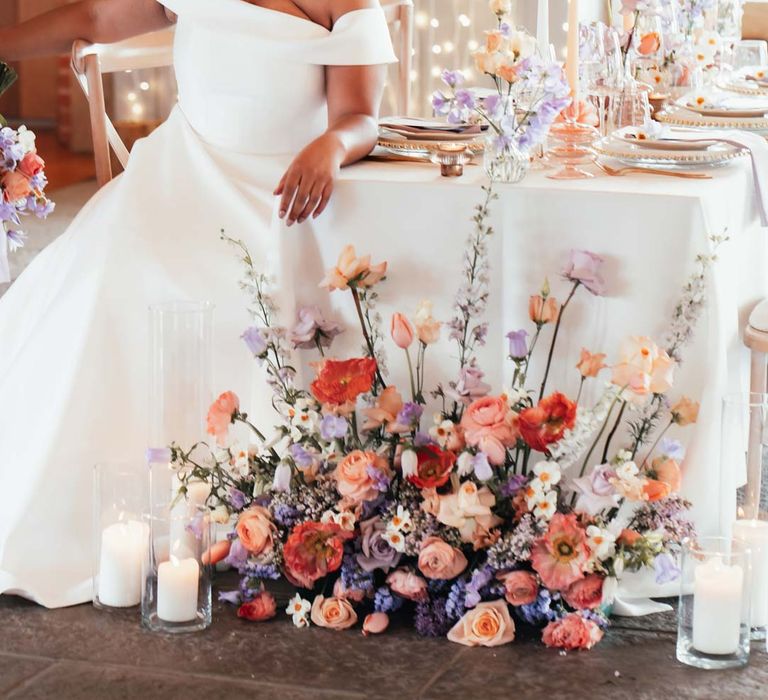 Bride in off the shoulder wedding dress with puddle train sitting at peach and lilac wedding tablescape with spring flower centrepieces, lilac napkins, tinted glassware and gold cutlery and crockery on white tablecloth with flower arrangement with peach garden roses, white peonies, lilac sweet peas, bluebells and wildflowers at Rackleys Barn