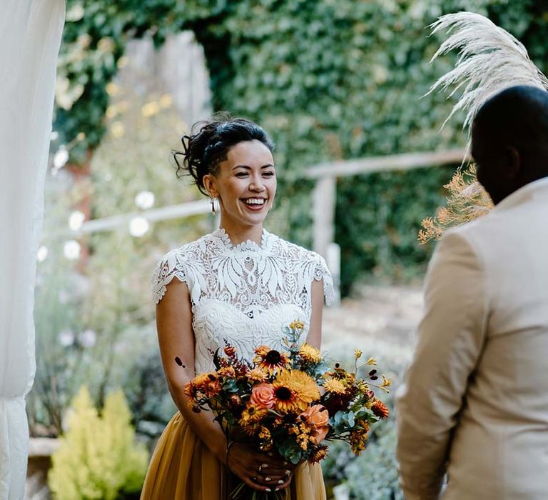 Bride in white mesh lace bridal crop top and yellow wedding skirt holding dried autumnal wedding bouquet with sunflowers, garden roses, pampas grass, eucalyptus, yellow tall kangaroo paws, orange osmanthus flowers, foliage and dried flowers and smiling at groom at Broadfield Court