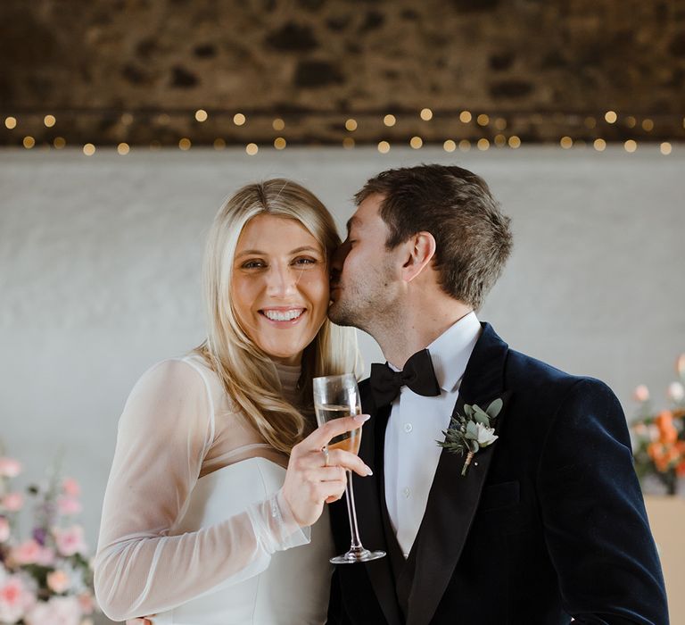 Groom in black tie kisses the bride on the cheek who holds a champagne glass