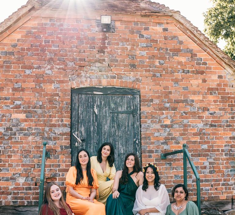 Bride in long sleeve mesh overlay wedding dress and colourful bridal flower crown sitting with bridesmaids in mismatched bridesmaid dresses at Templars Barn 