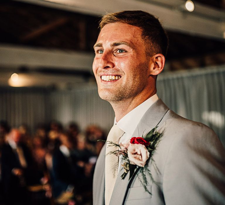 Groom in a light grey wedding suit with red and pink buttonhole flowers smiling standing at the altar 