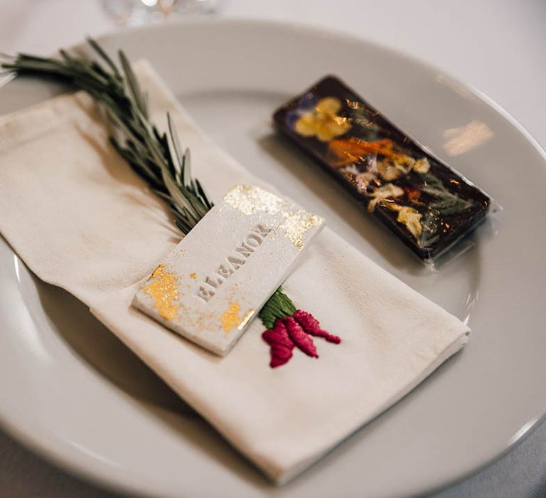 Wedding place setting with white plated decorated with embroidered wedding napkin, sprig of rosemary, and gold flake wedding place name 