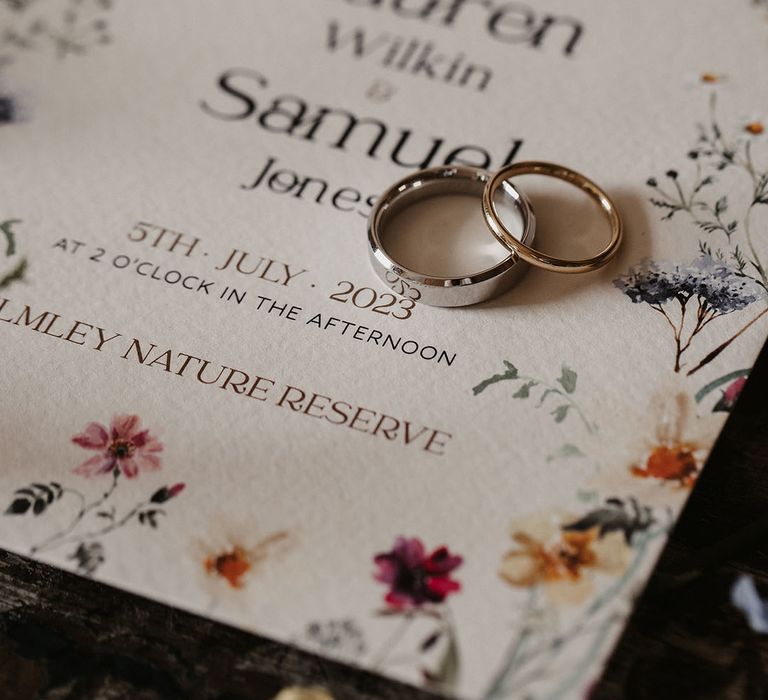 Two platinum wedding bands for the bride and groom resting on top of the floral wedding invitations 