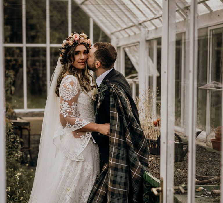 Groom in tartan kisses boho bride in rose flower crown in greenhouse at highland wedding in Glencoe