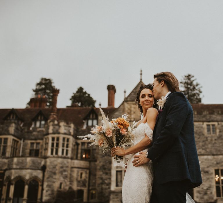 Groom in a navy suit kissing his brides forehead outside their Rhinefield House wedding venue. The bride wears a lace wedding dress and holds a dried flower bouquet 