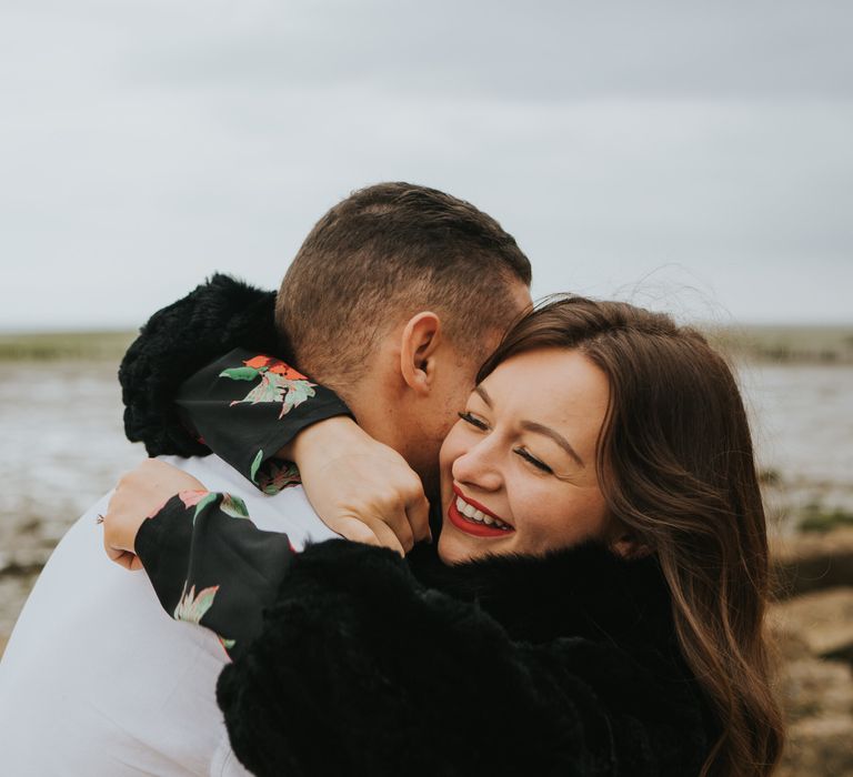 White brunette bride-to-be in a black faux fur coat wrapping her arms around her groom-to-be during the beach engagement photo shoot