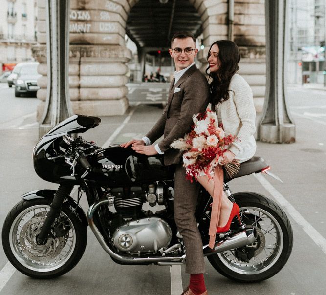 A newly wed couple sit on a motorbike at their Parisian elopement. The bride holds a bright red bouquet and red shoes. The groom wears a brown suit. Photography by Ann-Sophie Benoit.