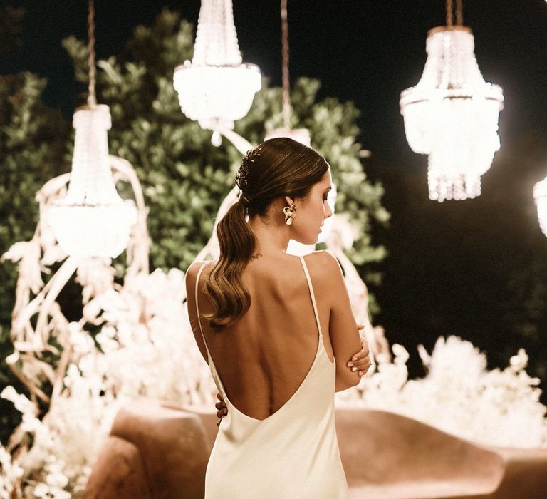 Brunette bride with a sleek ponytail in a low back satin slip dress standing by a chandelier light instalment. 