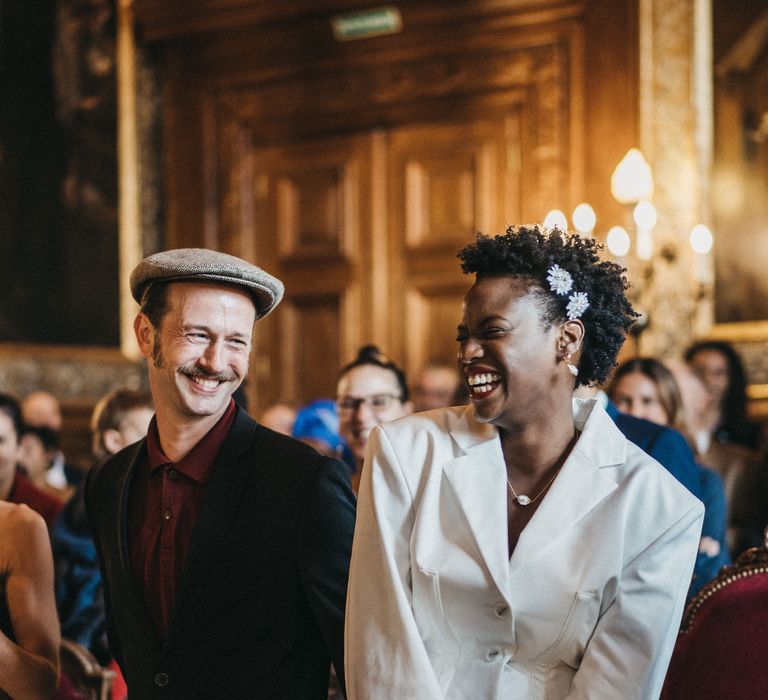 Groom in a Peaky Blinders wedding suit and flat cap smiling at the altar with his bride in a short blazer dress