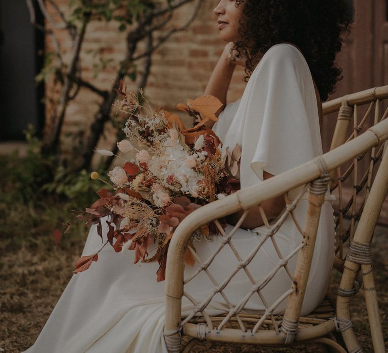 Beautiful bride in a crepe wedding dress with naturally curly hair sitting on a wicker chair holding an autumn leave wedding bouquet 