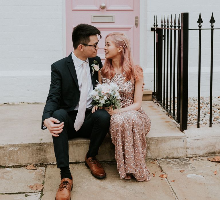 Bride in a pink sequin dress sitting on a step with her groom in a dark suit 