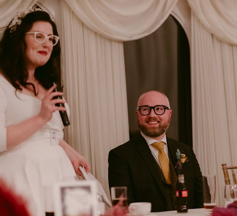 Smiling groom in navy suit and yellow tie smiles at bride in white Rime Arodaky dress and bridal headband as she makes speech