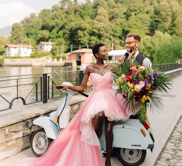 Black bride sits on pale blue vespa whilst wearing pastel pink wedding gown as groom stands behind her wearing yellow sunglasses