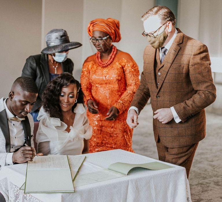 Groom in grey Moss Bros suit sits with bride in white Bec + Bridge wedding dress signing the marriage registry whilst wedding guests act as witnesses 