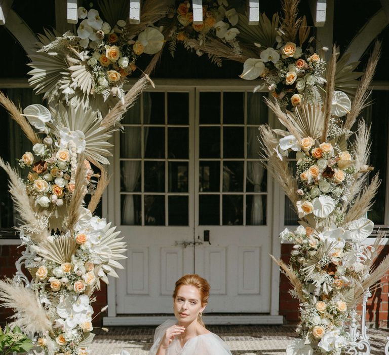 Bride sitting under boho inspired flower arch wearing a floral lace wedding dress with tulle shoulder detail