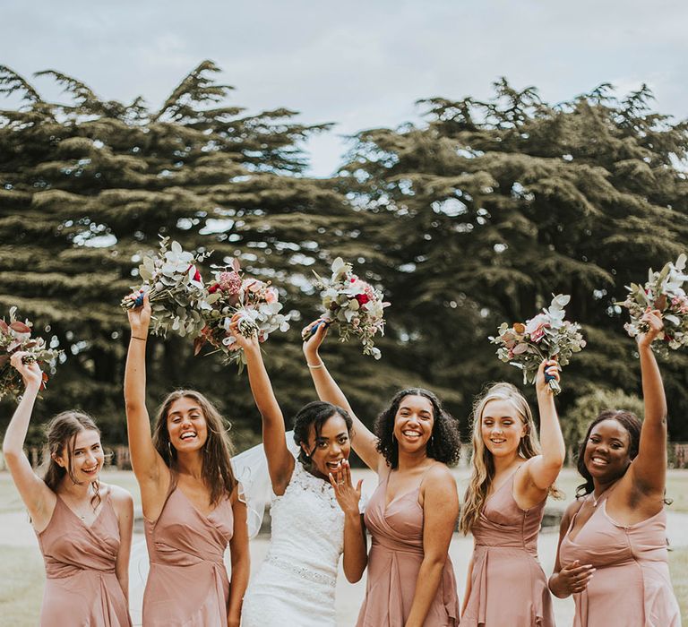 Fun bridal party portrait with bride and bridesmaids in dusky pink dresses holding their bouquets in the air 